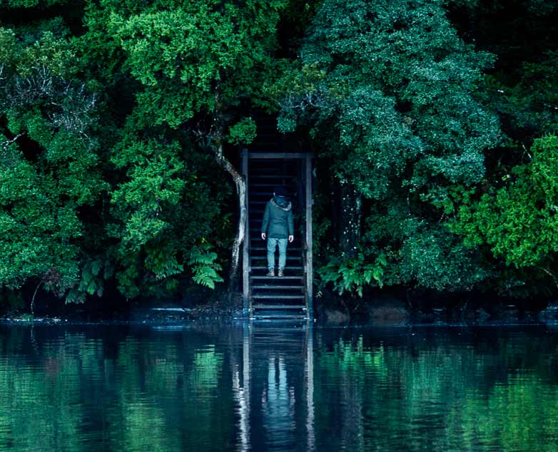 A man stands at the bottoms of some stairs that lead up into a dense forest, next to the calm waters of a river.