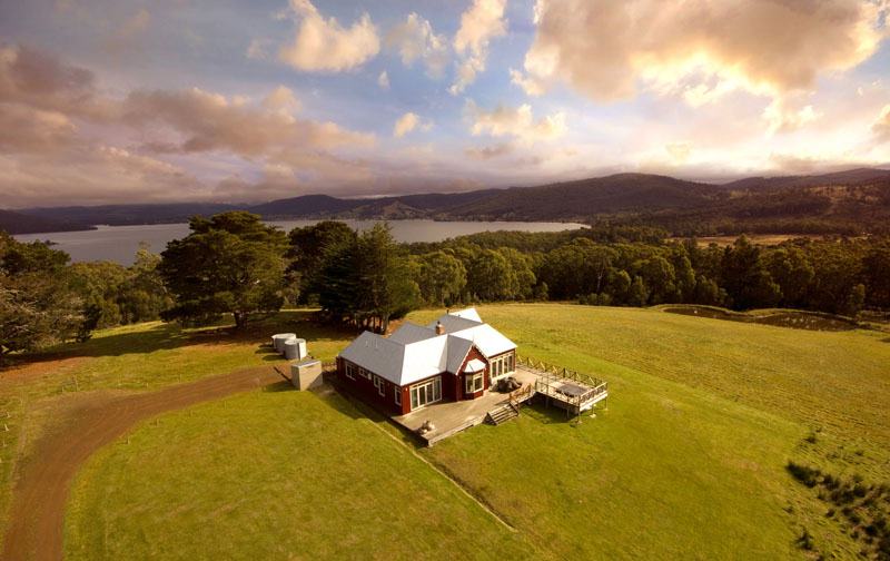A large house sits on top of a green grass hill surrounded by forest, with a river and mountain in the background