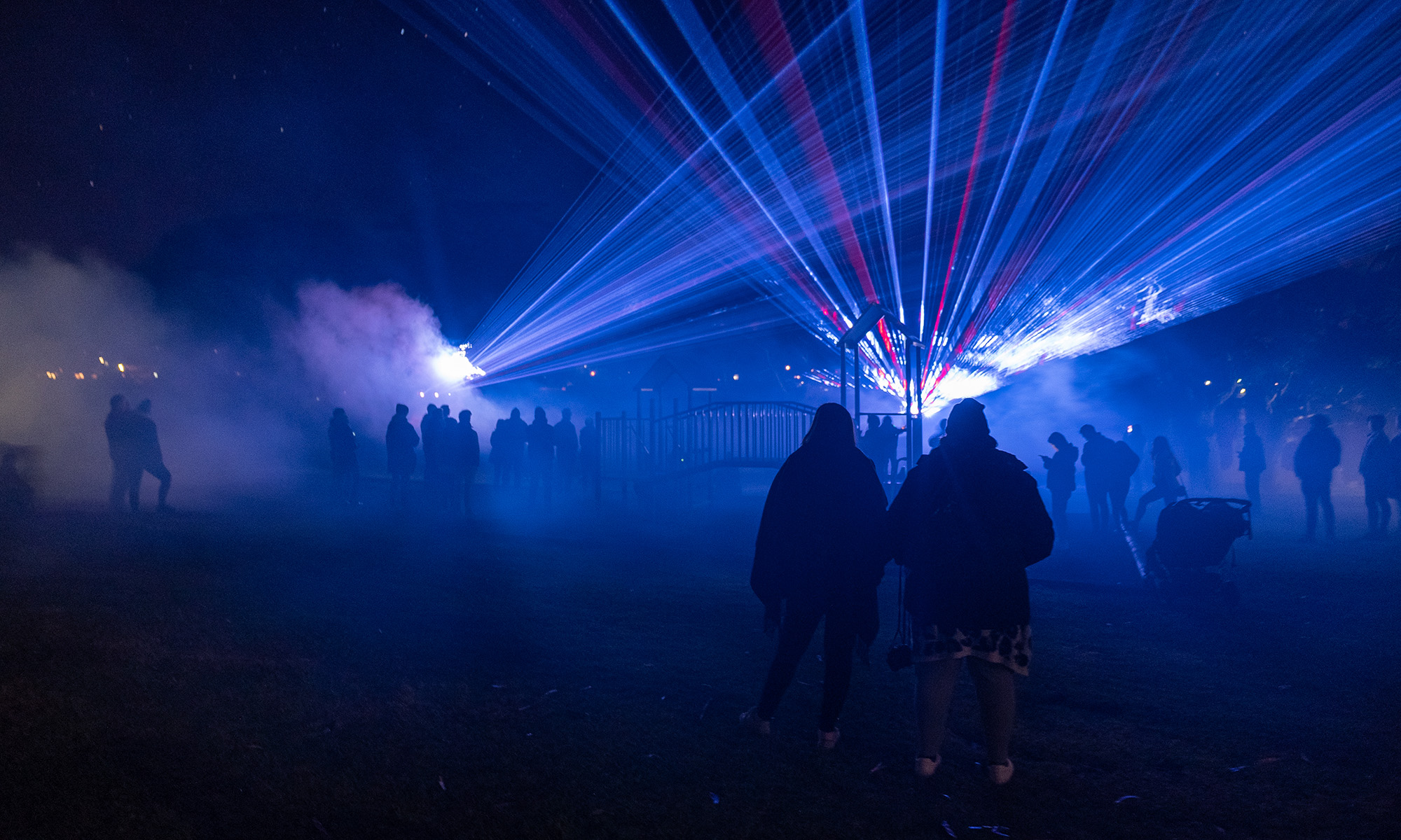Silhouettes of people standing among a foggy playground at night while beams of light shoot into the sky 