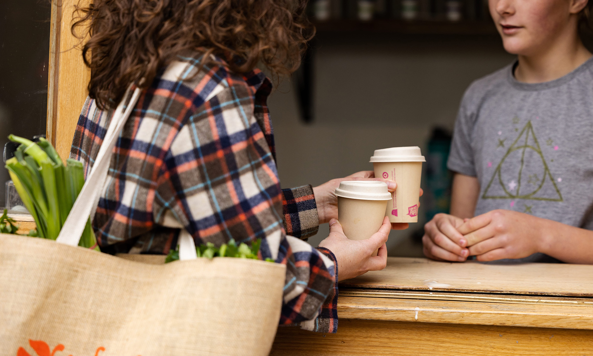 Over a wooden counter, a person carrying a tote bag of vegetables buys two takeaway coffees.