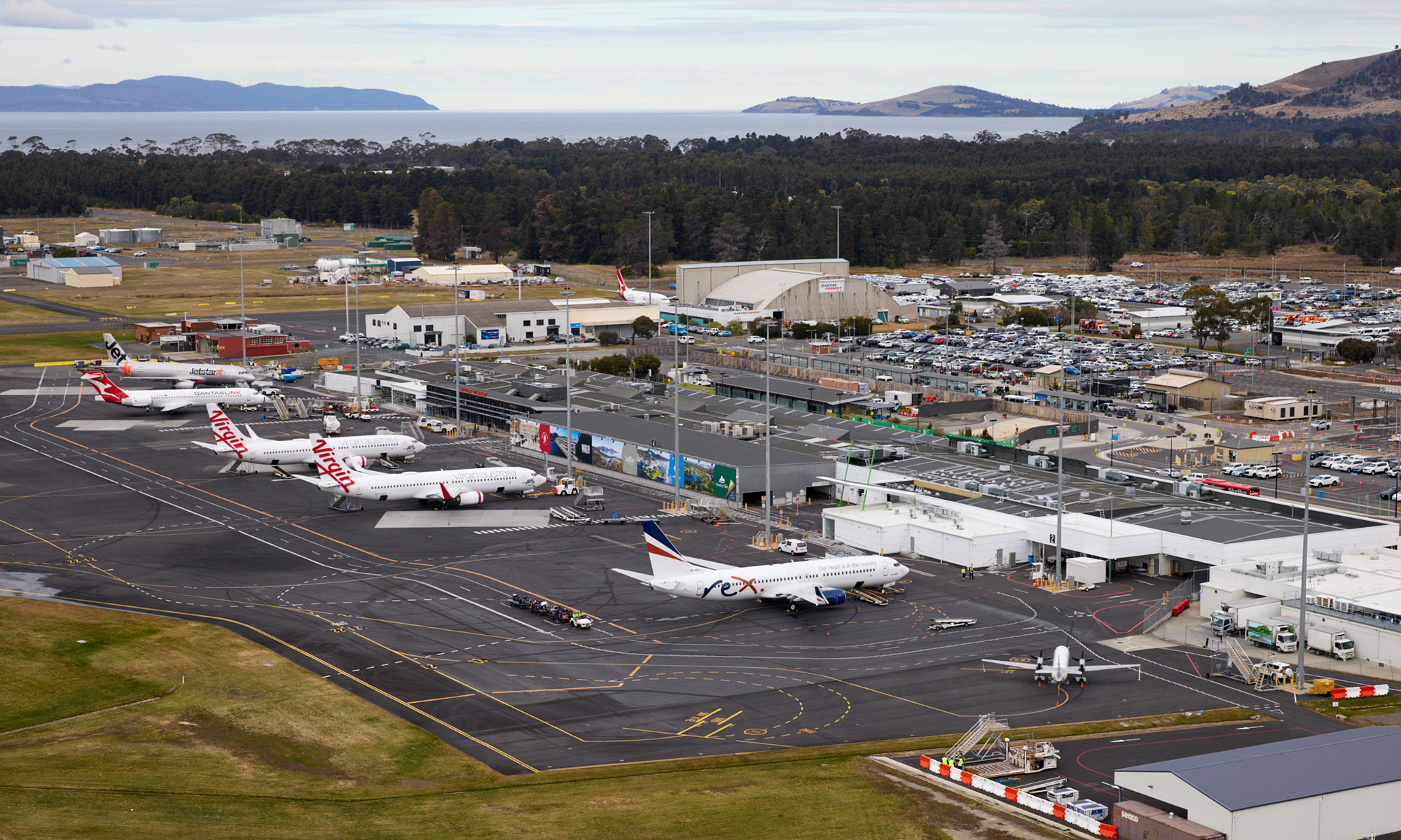 An aerial view of a small airport, with several planes lined up at the terminal. A body of water and hills rise up from past the treeline.